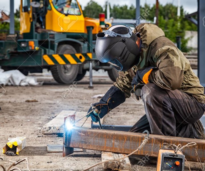 A young  man welder in brown uniform, welding mask and welders leathers, weld  metal  with a arc welding machine at the construction site, blue sparks fly to the sides